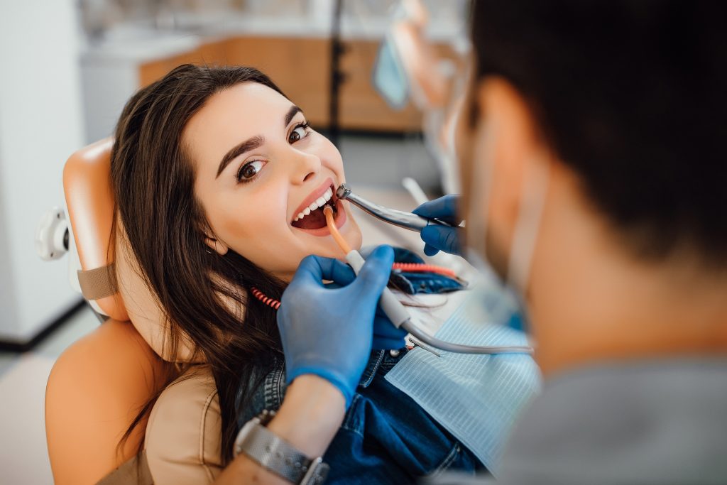 Young female patient visiting dentist office. Beautiful woman with healthy straight white teeth sitting at dental chair with open mouth during oral checkup while doctor working at teeth.